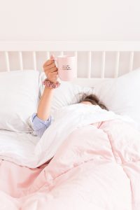 Woman in bed, covered by the duvet, holding up a mug which reads 'I am a lot cooler on the internet'