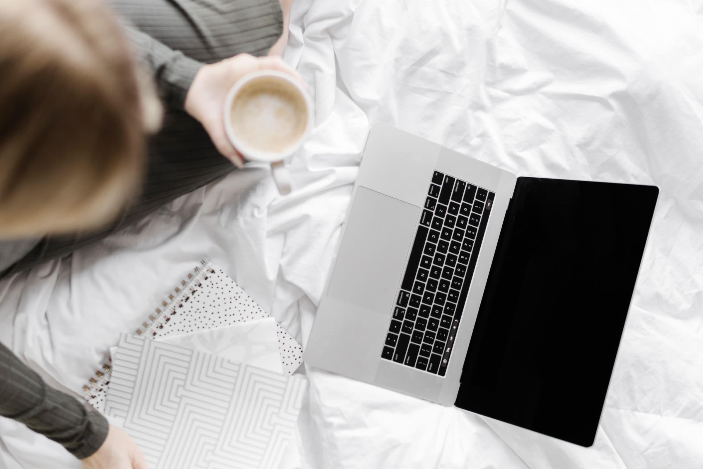 Woman sat on a bed with a cup of tea in her hand and a laptop on the bed