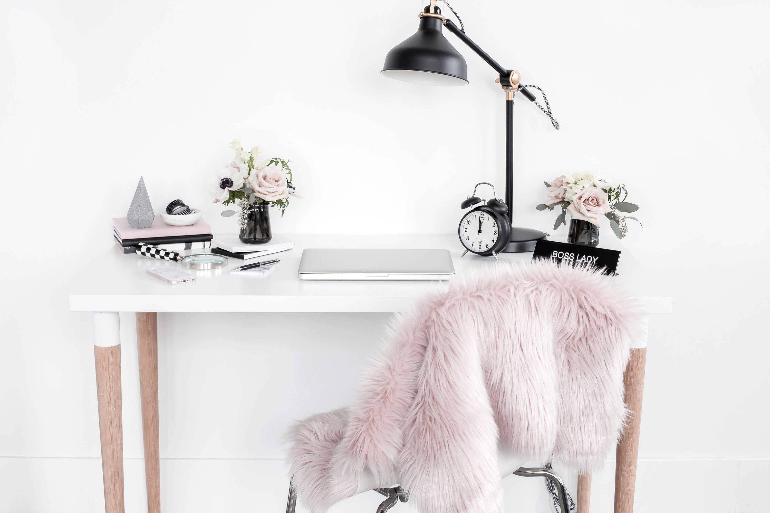 White desk with chair. The chair is covered in a pink fluffy throw. The desk has various items including a laptop, a desk lamp and an alarm clock