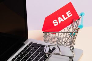 Tiny shopping trolley / cart sitting on a laptop. Inside the cart there is a large red label with the word SALE written on it in white