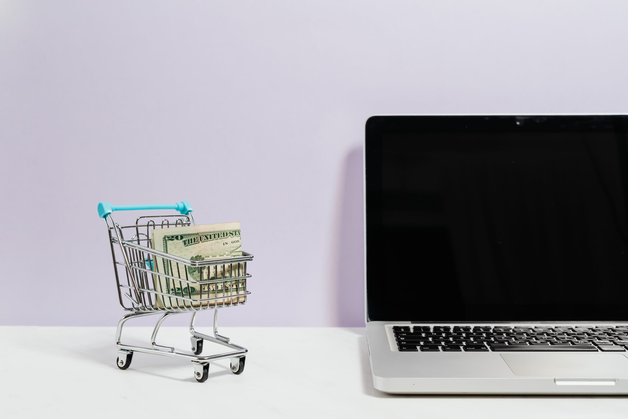 Tiny shopping cart filled with banknotes next to a laptop